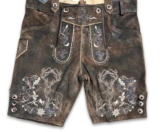 Könnte beinhalten: Dunkelbraune Lederhose mit detaillierter weißer Stickerei, die Hirsche, florale Motive und Bergsilhouetten zeigt. Die Hose ist mit Hosenträgern, seitlichen Metallknöpfen am Bund und einem kleinen angebrachten Kunststoffetikett versehen.