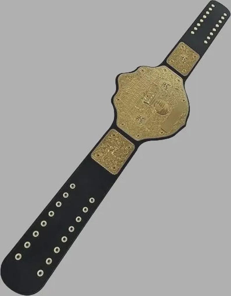 Puede incluir: Un cintur&oacute;n de campeonato con una correa de cuero negro, ojales met&aacute;licos plateados y tres placas de metal dorado. La placa central es grande y octogonal con grabados intrincados, mientras que las dos placas laterales son cuadradas.