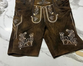 Könnte beinhalten: Dunkelbraune Lederhose aus Wildleder mit cremefarbener Stickerei von Hirschen und floralen Motiven auf den Vorderteilen. Die Hose verfügt über einen Latz, Seitentaschen und eine Lederschnürung am hinteren Bund zur individuellen Anpassung.
