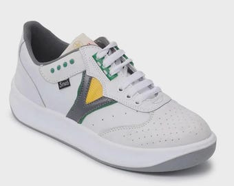 Könnte beinhalten: Ein weißer Sneaker mit perforierter Zehenkappe, weißen Schnürsenkeln und grauem Innenfutter. Die Seite zeigt einen grauen V-förmigen Besatz, einen gelben dreieckigen Akzent, grüne Ösen und ein schwarzes Etikett mit dem weißen Text Sovis.