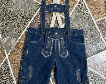 Könnte beinhalten: Dunkelblaue Jeans-Lederhosen-Shorts mit heller Zierstickerei auf dem Latz und den Beinen. Die Shorts verfügen über verstellbare Hosenträger mit Metallschnallen, einen Latzverschluss und drei kleine Knöpfe an jedem äußeren Beinabschluss.