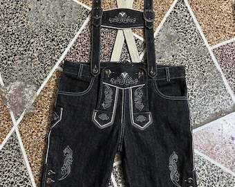 Puede incluir: Pantalones cortos tipo lederhosen de mezclilla gris oscuro con costuras blancas en contraste, bordados intrincados en la solapa frontal y las piernas, tirantes ajustables con hebillas metálicas y tres pequeños botones metálicos en cada dobladillo.