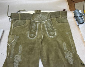 Könnte beinhalten: Olivgrüne Lederhose mit aufwendigen weißen Stickereien. Die Hose hat eine Latzfront mit dekorativen Nähten und Taschen. Das Kleidungsstück hat ein klassisches Design mit Gürtelschlaufe und Seitenverschluss.