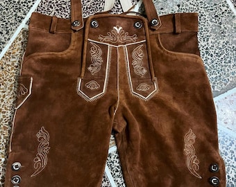 Könnte beinhalten: Braune Lederhose aus Wildleder mit detaillierten cremefarbenen Stickereien auf dem Latz und den Beinen. Das Kleidungsstück verfügt über verstellbare Träger mit Metallschnallen, einen Latzverschluss vorne und drei Zierknöpfe an jedem Beinabschluss.