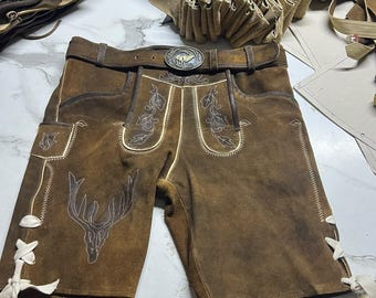Könnte beinhalten: Braune Lederhose mit weißen Ziernähten, einer Hirschkopf-Stickerei auf dem linken Bein und seitlicher Schnürung mit weißen Bändern. Die Hose enthält einen passenden Gürtel mit einer runden Metallschnalle, die ein Hirschmotiv zeigt.