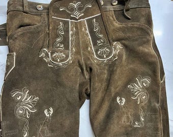 Könnte beinhalten: Dunkelbraune Lederhose aus Wildleder mit einem Latz, der eine aufwendige cremefarbene Stickerei aufweist. Das Design umfasst florale Verzierungen und stilisierte Hirschmotive auf beiden Beinen sowie cremefarbene Paspeln entlang der Nähte.