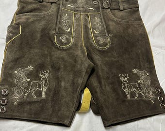Könnte beinhalten: Dunkelbraune Lederhose aus Wildleder mit hellgelben Paspeln und cremefarbener Stickerei. Das Design zeigt detaillierte Hirschmotive an den Beinen, florale Muster auf dem Latz sowie metallische Knöpfe an den Seiten der kurzen Hose.