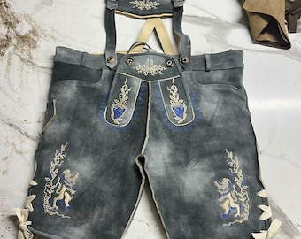 Könnte beinhalten: Dunkelgraue Lederhose mit hellen Trägern, aufwendigen cremefarbenen und blauen Stickereien auf dem Latz und den Beinen sowie seitlichen Schnürungen. Das Kleidungsstück verfügt über einen Brustlatz mit passender Stickerei und Metallschnallen.