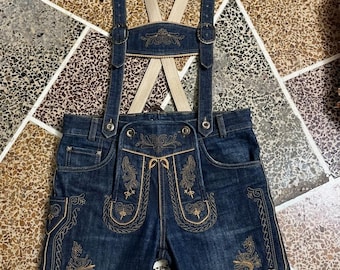 Könnte beinhalten: Dunkelblaue Lederhosen-Shorts aus Denim mit hellbeiger Stickerei auf den Vorderteilen und Beinabschlüssen. Das Kleidungsstück verfügt über verstellbare beige Hosenträger mit passenden Denim-Querriemen und Metallknöpfen an den Seiten.