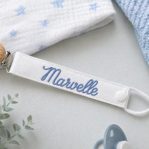 Peut inclure: Une attache-sucette en tissu blanc avec le pr&eacute;nom Marvelle brod&eacute; en &eacute;criture cursive bleue. La sangle comprend une pince en m&eacute;tal argent&eacute; &agrave; une extr&eacute;mit&eacute; et un bouton-pression blanc avec une boucle &eacute;lastique blanche pour fixer la sucette.