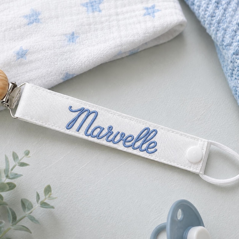 Peut inclure: Une attache-sucette en tissu blanc avec le pr&eacute;nom Marvelle brod&eacute; en &eacute;criture cursive bleue. La sangle comprend une pince en m&eacute;tal argent&eacute; &agrave; une extr&eacute;mit&eacute; et un bouton-pression blanc avec une boucle &eacute;lastique blanche pour fixer la sucette.