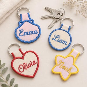 May include: Four embroidered fabric keychains with metal rings. One pink shell shape with blue trim and the name Emma, one cream circle with blue trim and the name Liam, one cream heart with red trim and the name Olivia, and one pink star with yellow trim and the name Noah.