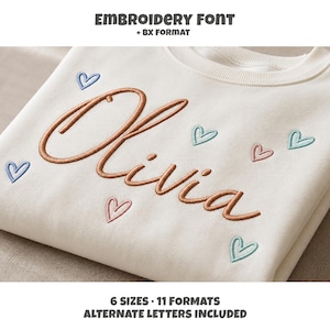 May include: An off-white sweatshirt featuring the embroidered name Olivia in a brown cursive script, surrounded by small embroidered hearts in blue, pink, and teal. Text at the top reads EMBROIDERY FONT + BX FORMAT and at the bottom 6 SIZES 11 FORMATS ALTERNATE LETTERS INCLUDED.