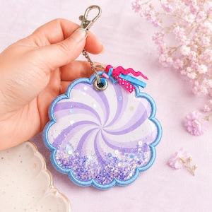 May include: A circular scalloped keychain featuring a purple and white spiral pattern with a clear window filled with purple and blue glitter. The edge is finished with blue embroidery, and it includes a silver lobster clasp and small ribbons.