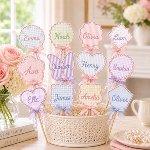 May include: A collection of personalized decorative sticks featuring various shapes like hearts, flowers, and butterflies with gingham patterns. Each stick displays a name embroidered in cursive, including Emma, Noah, Olivia, Liam, Ava, Charlotte, Henry, Sophia, Ella, James, Amelia, and Oliver, accented with small satin bows.
