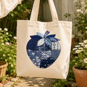 May include: A cream-colored canvas tote bag featuring a large, central strawberry-shaped appliqué. The appliqué is constructed from various shades of blue fabric patches, showcasing diverse patterns including hearts, stars, and floral motifs.