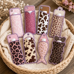 May include: Eight fabric lip balm holders with various patterns including pink leopard print, dark pink glitter, brown and white cow print, and tan leopard print. Each holder features a small circular cutout at the top and a white tube inside.