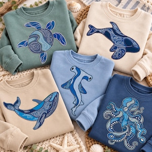 May include: Five crewneck sweatshirts in various colors, each featuring a detailed embroidered marine animal design. The collection includes a sea turtle, an orca, a hammerhead shark, a whale, and an octopus, all rendered in shades of blue.