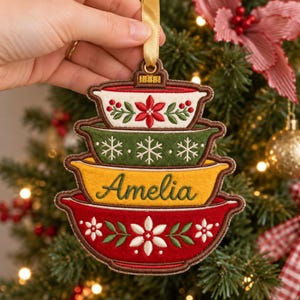 May include: A felt Christmas tree ornament shaped like a stack of four colorful mixing bowls. The top bowl is white with red poinsettias, the second is green with white snowflakes, the third is yellow with the name Amelia, and the bottom is red.