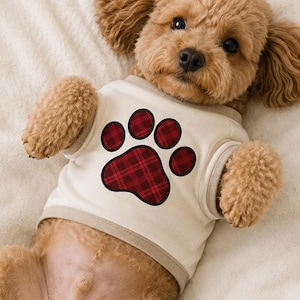 May include: A light beige pet shirt featuring a large paw print graphic on the chest. The paw print is filled with a red and black plaid pattern and outlined in black. The shirt has a ribbed crew neckline and arm openings in a matching beige tone.