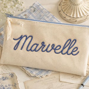 Peut inclure: Une trousse rectangulaire en toile blanc cass&eacute; avec le nom Marvelle brod&eacute; en &eacute;criture cursive bleue. La trousse comporte une fermeture &eacute;clair bleu clair avec un petit ruban de dentelle cr&egrave;me attach&eacute; au curseur et une petite &eacute;tiquette lat&eacute;rale.