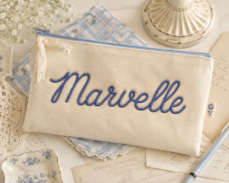 Peut inclure: Une trousse rectangulaire en toile blanc cass&eacute; avec le nom Marvelle brod&eacute; en &eacute;criture cursive bleue. La trousse comporte une fermeture &eacute;clair bleu clair avec un petit ruban de dentelle cr&egrave;me attach&eacute; au curseur et une petite &eacute;tiquette lat&eacute;rale.
