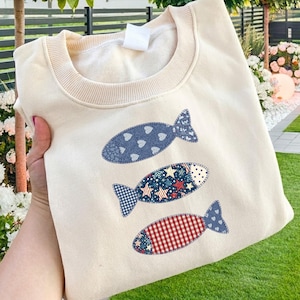 May include: A cream-colored crewneck sweatshirt featuring three fish-shaped appliques arranged vertically. The top fish is blue with white hearts, the middle fish has a red, white, and blue star pattern, and the bottom fish features a red gingham check.