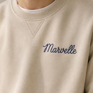 Peut inclure: Un sweat-shirt beige &agrave; col rond avec un d&eacute;tail de couture en V au niveau de l'encolure. Le mot Marvelle est brod&eacute; en &eacute;criture cursive bleu fonc&eacute; sur la partie sup&eacute;rieure gauche de la poitrine du v&ecirc;tement, port&eacute; sur un t-shirt blanc.