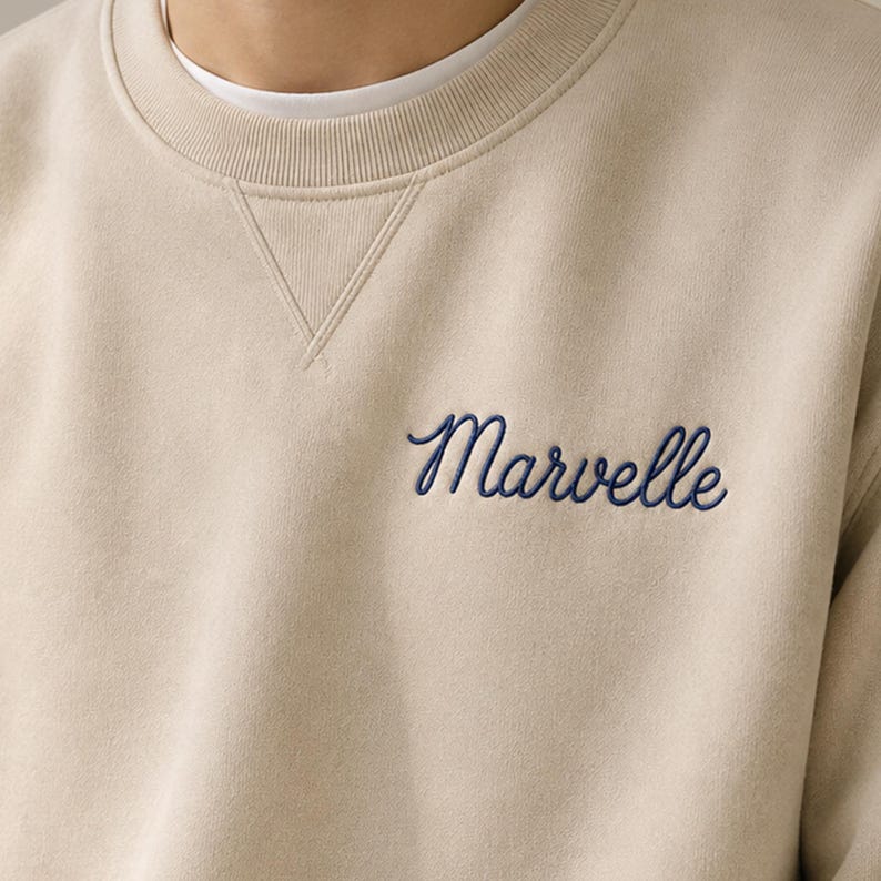 Peut inclure: Un sweat-shirt beige &agrave; col rond avec un d&eacute;tail de couture en V au niveau de l'encolure. Le mot Marvelle est brod&eacute; en &eacute;criture cursive bleu fonc&eacute; sur la partie sup&eacute;rieure gauche de la poitrine du v&ecirc;tement, port&eacute; sur un t-shirt blanc.