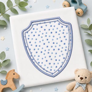 May include: A white fabric square featuring a centered shield-shaped applique. The shield is white with a pattern of small blue polka dots and is outlined with a thick, stitched blue border. The design is clean, centered, and clearly defined.