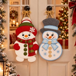 May include: Two felt snowman wall hangings suspended by gold cords on a double door. The left snowman wears a red hat and plaid scarf. The right snowman wears a gray top hat and a light blue bow tie with snowflake embroidery and gold buttons.