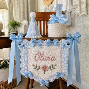 May include: A first birthday party set featuring a light blue gingham party hat with a white pom-pom, a cake topper with the number one, and a rectangular fabric banner with scalloped edges. The banner displays the name Olivia in pink script.