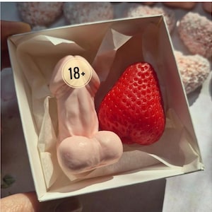 May include: A white gift box containing a pale pink phallic-shaped object and a bright red strawberry-shaped item. The pink object features a circular sticker with the text 18+ printed in black. The items are nestled inside white tissue paper.