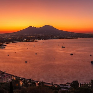 Puede incluir: Una vista panorámica de la bahía de Nápoles al atardecer, con la silueta del monte Vesubio sobre un cielo vibrante de color naranja y púrpura. La costa está iluminada por las luces de la ciudad y hay varios barcos pequeños en el agua.