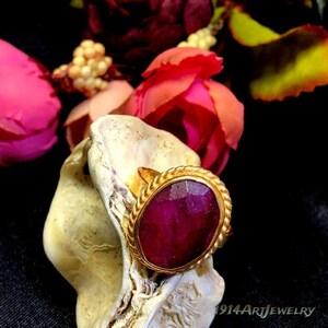 Op de afbeelding: Een gouden ring met een grote, ovaalvormige, gefacetteerde donkerroze edelsteen in een gedraaide, touwachtige metalen band. De ring is geplaatst op een getextureerde, gebroken witte schelp. De tekst 1914ArtJewelry is zichtbaar in de rechterbenedenhoek.