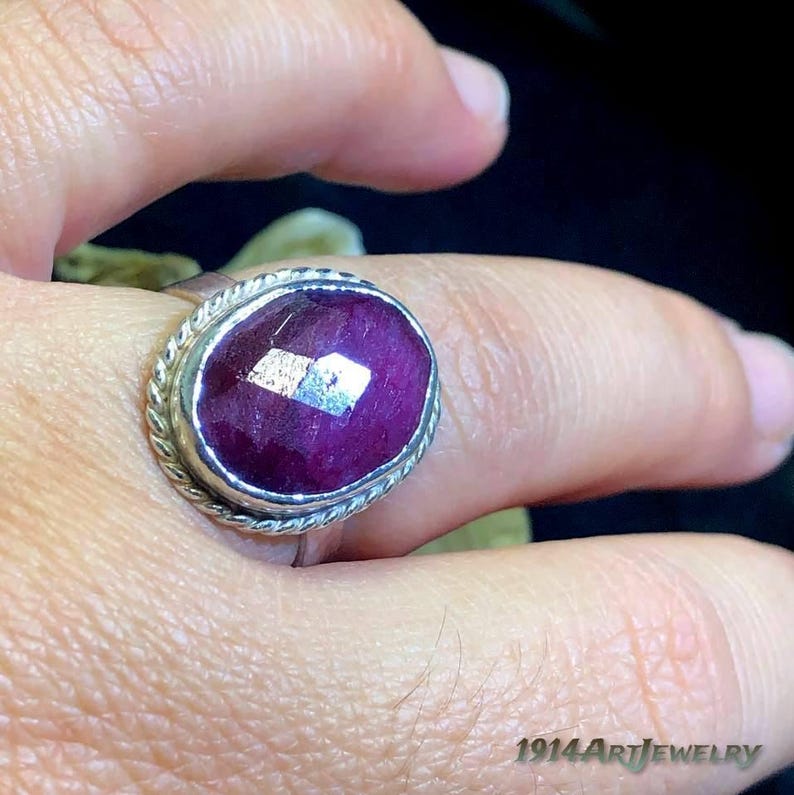 Op de afbeelding: Een ring met een grote, ovaalvormige, gefacetteerde diepmagenta edelsteen in een zilveren zetting. De zetting is omringd door een gedraaide, touwachtige metalen rand. De tekst 1914ArtJewelry is zichtbaar in de rechterbenedenhoek van de afbeelding.