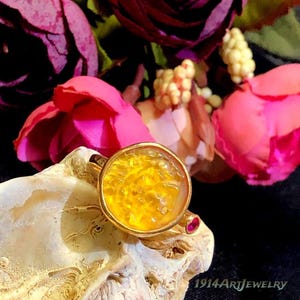 May include: A gold ring featuring a round, textured yellow amber-colored stone in a bezel setting, accented by a small, round, vibrant pink gemstone on the side of the band, with the text 1914ArtJewelry visible at the bottom right corner.