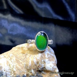May include: A silver ring featuring a central oval-shaped mood stone that displays a vibrant green color. The ring band is accented with a small, clear, round stone on each side of the central setting. The text 1914ArtJewelry is visible.