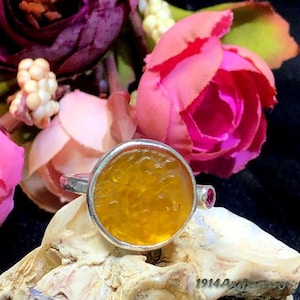 May include: A silver ring featuring a circular, textured amber-colored stone set in a smooth metal bezel. The ring band includes a small, round, deep pink gemstone accent on the side, providing a contrast to the central yellow stone.