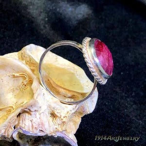 Op de afbeelding: Een zilveren ring met een ronde, gefacetteerde, dieproze-rode edelsteen gezet in een gedraaide, touwachtige metalen zetting. De ring ligt op een getextureerde, gebroken witte schelp. De tekst 1914ArtJewelry is rechtsonder zichtbaar.