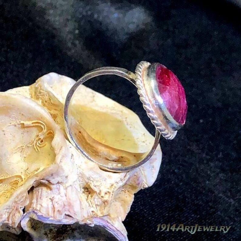 Op de afbeelding: Een zilveren ring met een ronde, gefacetteerde, dieproze-rode edelsteen gezet in een gedraaide, touwachtige metalen zetting. De ring ligt op een getextureerde, gebroken witte schelp. De tekst 1914ArtJewelry is rechtsonder zichtbaar.