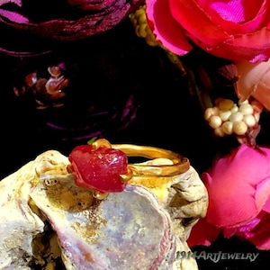 May include: A gold ring featuring a raw, deep red gemstone set in a prong setting. The band has a textured, organic finish. The text 1914ArtJewelry is visible in the bottom right corner of the image, set against a dark background with flowers.