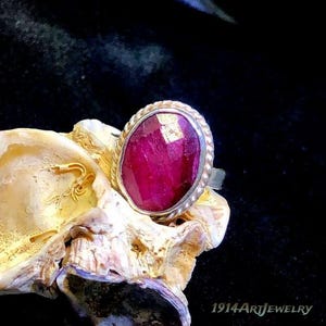 Op de afbeelding: Een ring met een grote, ovaalvormige, dieproze gefacetteerde edelsteen in een zilverkleurige metalen band met een gedraaide touwrand. De ring rust op een getextureerde, cr&egrave;mekleurige schelp. De tekst 1914ArtJewelry is zichtbaar op de afbeelding.