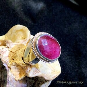 Op de afbeelding: Een zilveren ring met een grote, ovaalvormige, gefacetteerde donkerroze edelsteen in een zetting met een gedraaide touwrand. De ringband is glad en metaalachtig. De tekst 1914ArtJewelry is zichtbaar in de rechterbenedenhoek.
