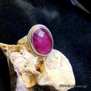 Op de afbeelding: Een ring met een grote, ovaalvormige, gefacetteerde donkerroze edelsteen in een zilverkleurige metalen band met een gedraaide koordrand. De ring ligt op een gestructureerd, lichtgekleurd mineraaloppervlak met de tekst 1914ArtJewelry.