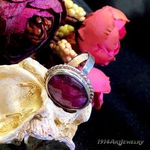 Op de afbeelding: Een zilveren ring met een grote, ovale, diepmagenta edelsteen met een gefacetteerd oppervlak, omringd door een gedraaide metalen rand. De ring is geplaatst tegen een achtergrond van donkerroze bloemen en een getextureerde, cr&egrave;mekleurige schelp.