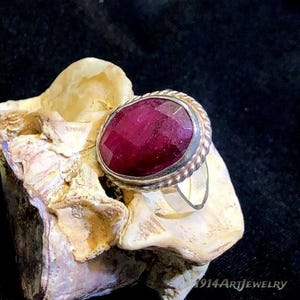Op de afbeelding: Een ring met een grote, ovaalvormige, diep magentarode edelsteen gezet in een zilverkleurige metalen band met een gedraaide, touwachtige rand. De ring is geplaatst op een textuurrijk, cr&egrave;mekleurig mineraal tegen een donkere achtergrond.