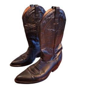Könnte beinhalten: Ein Paar schwarze Cowboystiefel aus Leder mit spitzer Zehenpartie, einem Blockabsatz und dekorativen, farblich abgestimmten Ziernähten am hohen Schaft. Die Stiefel haben Zuglaschen am oberen Rand und eine glatte Oberfläche mit sichtbaren Gehfalten.