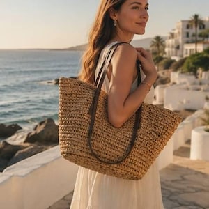 May include: A woven straw tote bag with brown leather handles is carried over the shoulder. The bag is rectangular and appears to be empty. The background shows a coastal scene with the ocean and buildings.
