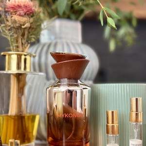 Może przedstawiać: Bursztynowa szklana butelka perfum o pojemności 100 ml 3,4 fl oz z brązową nakrętką i napisem MYKONOS, w towarzystwie dwóch małych szklanych fiolek z atomizerami w kolorze złotym, oznaczonych jako 3 ml i 5 ml, zawierających przezroczysty płyn.